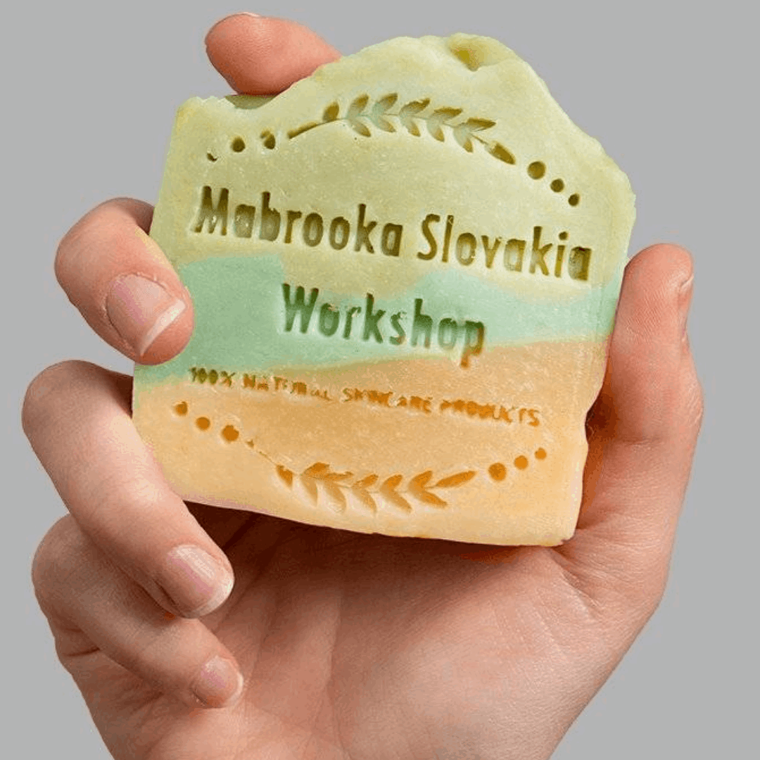 Luxurious bar soap made with cucumber slices, lemon slices, and aloe vera leaves on a wooden cutting board. Mabrooka logo in the background. hold by female hand 